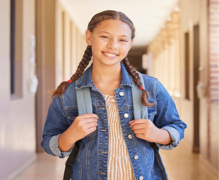 A young girl with braids smiling and holding a backpack in a school hallway.