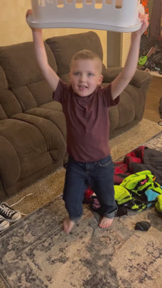 A young child joyfully lifting a laundry basket in a living room with clothes on the floor.