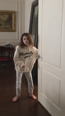 A young girl in a hoodie and pants standing near a door, looking into the room.